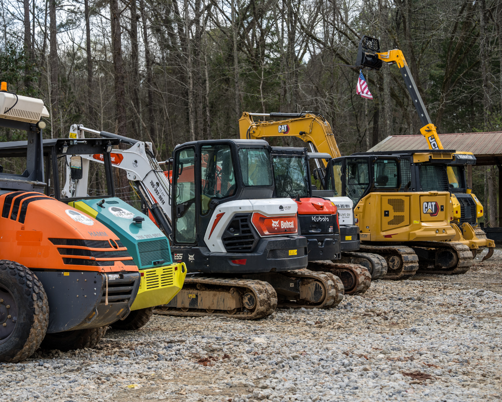 Row of construction excavators including Bobcat, Kubota, and CAT machines parked on gravel with trees in background