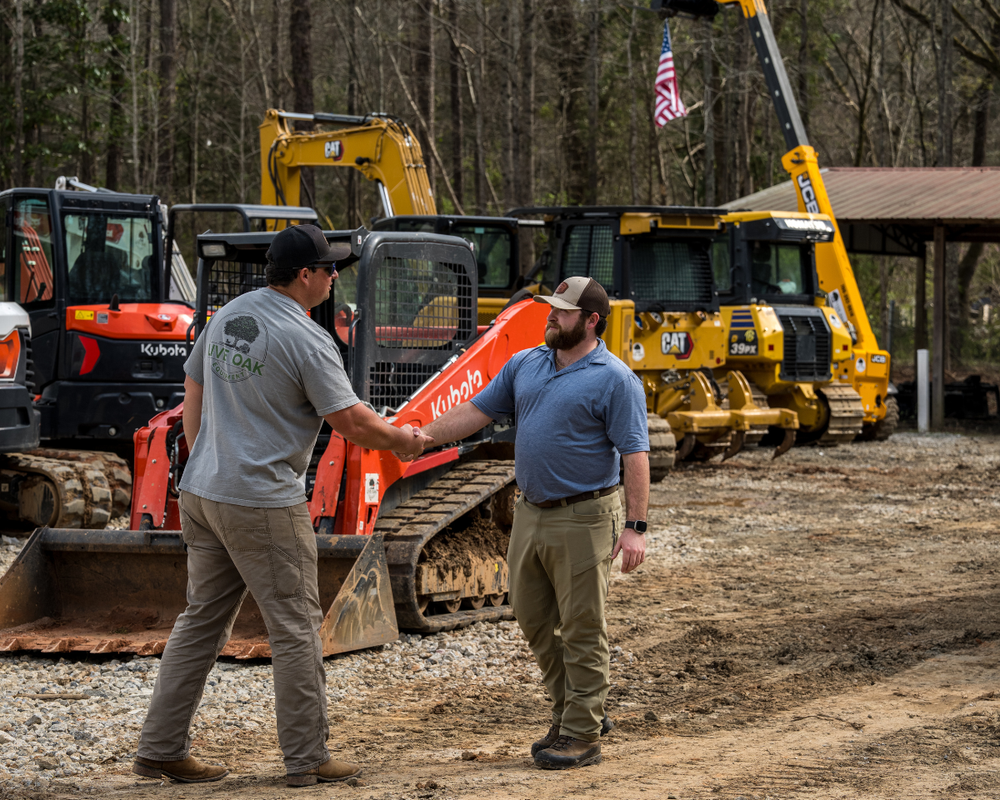 Two men shaking hands at an equipment dealership lot with Kubota, CAT, and JCB machinery behind them