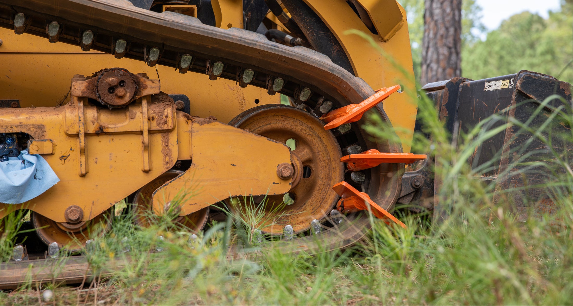 Close-up of yellow tracked machine with orange cutting teeth attachment in grassy forest setting