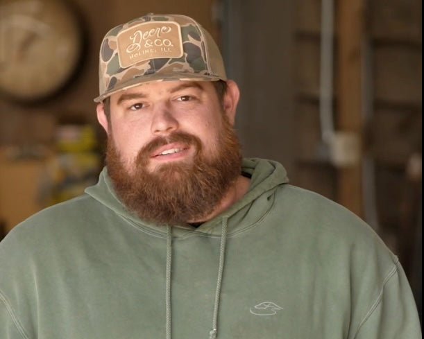 Man with red beard wearing green hoodie and camo "Deere & Co." trucker hat, indoors
