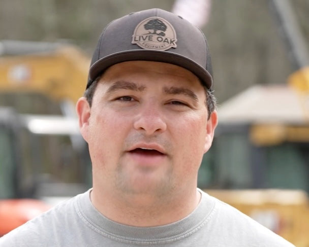 Man in brown "Live Oak Equipment" trucker cap and gray shirt, outdoors near construction equipment