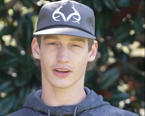 Young man in gray Realtree snapback cap and gray hoodie, outdoors with green foliage behind him