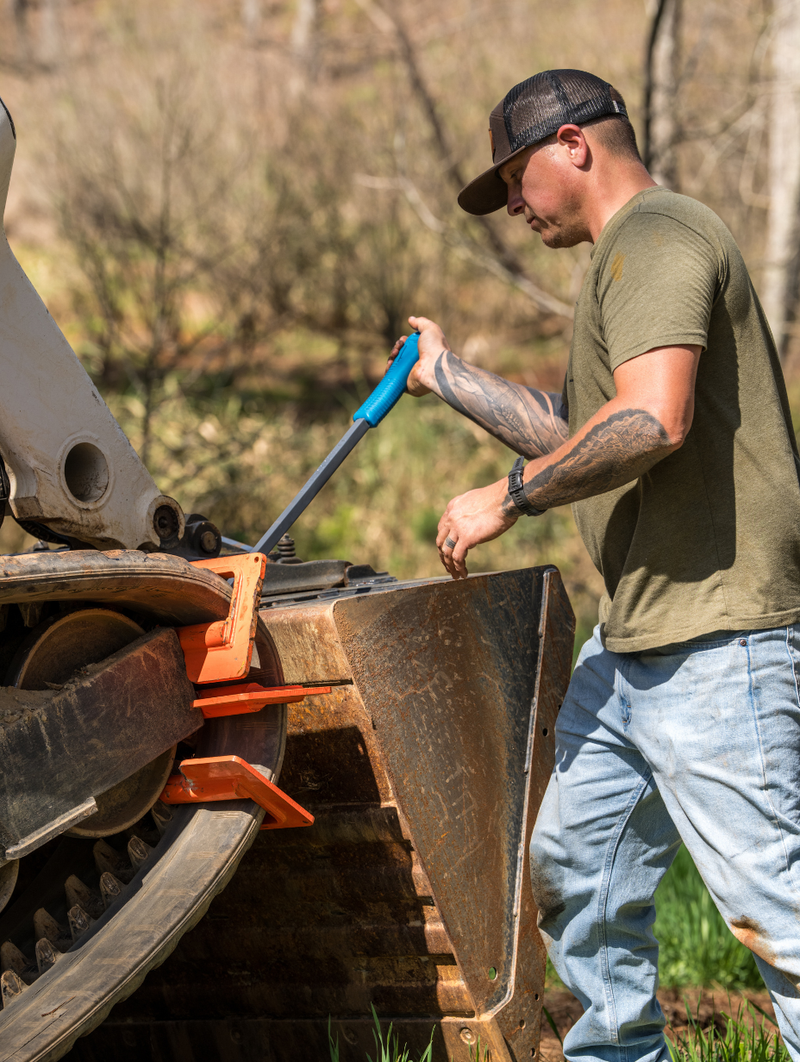 Tattooed man in green shirt uses blue pry bar on orange skid steer bucket outdoors