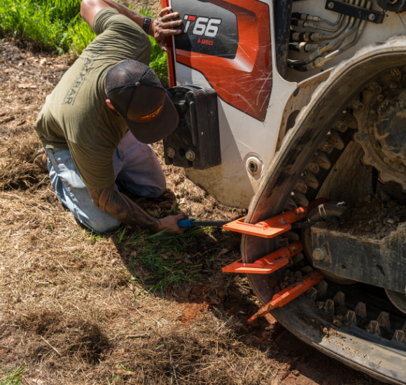 Man in green shirt and cap repairing orange track on a Bobcat T66 skid steer outdoors