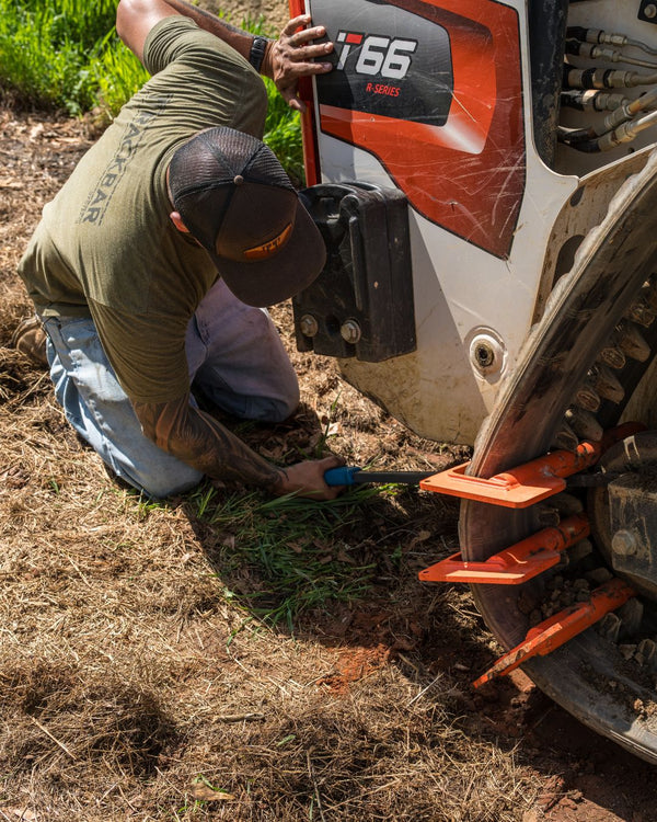 Technician in green shirt kneeling beside orange Bobcat T66 R-Series compact track loader, performing maintenance with a hand tool