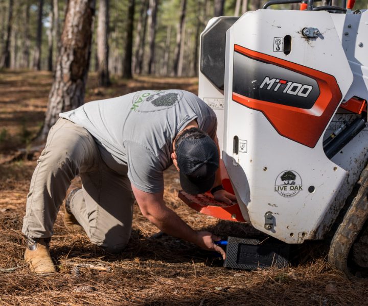 Man in gray shirt kneeling to service a white and orange Bobcat MT100 mini track loader in a pine forest