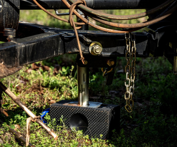 Black trailer jack resting on a rubber track block pad on green ground, with safety chains and hydraulic hoses visible above
